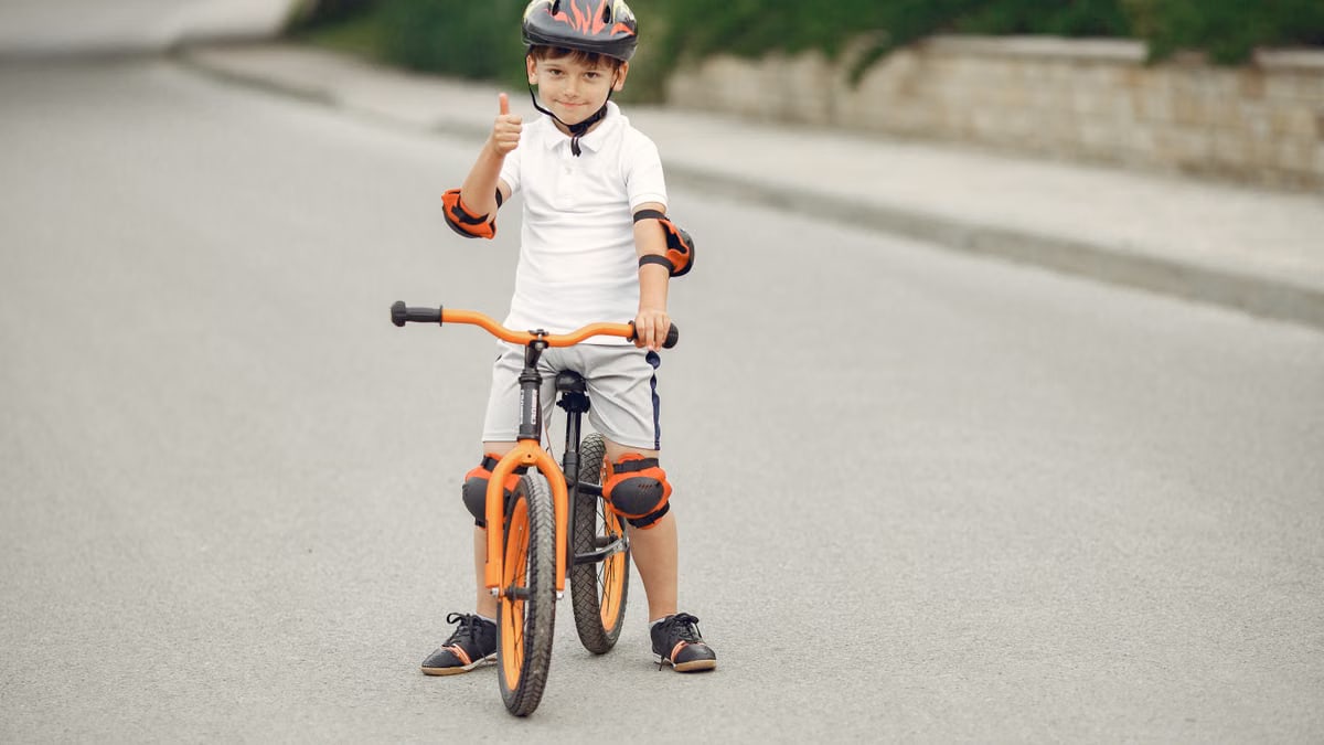 Smiling boy on an orange bike showing thumbs up on a sunny day, wearing safety gear.